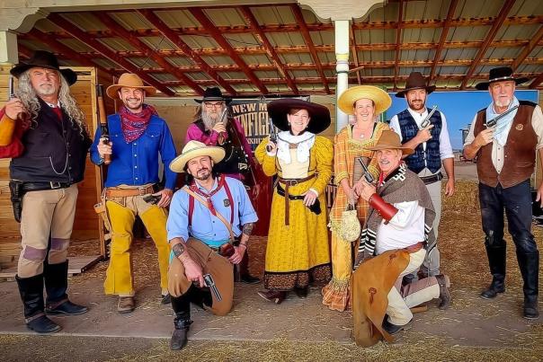 Group of people dressed as cowboys and cowgirls with guns posing under a wooden-roofed shelter in a western-themed setting.
