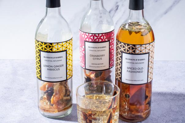 Three bottles of cocktail infusions labeled Lemon Ginger Hibiscus, Cranberry Citrus, and Spiced Old Fashioned stand on a marble surface, with a glass of infused drink in front.