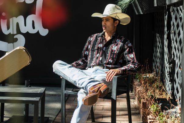 Dzaki Sukarno, New Mexico country artist, sitting outdoors in a cowboy hat and boots.