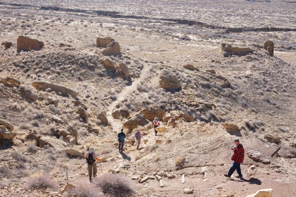 A group of people hike along a rugged trail in a vast desert landscape, surrounded by scattered rock formations.