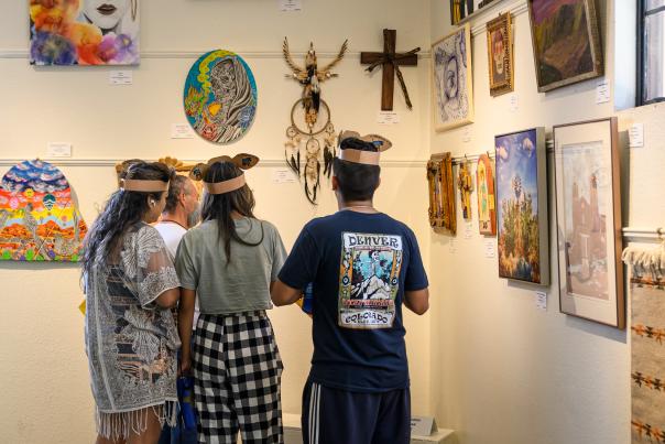 Four fair visitors view paintings, crafts, and mixed-media artworks on display inside an exhibit hall at the New Mexico State Fair.