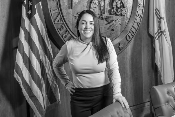 A person stands in a conference room beside a large carved Great Seal of the State of New Mexico, flanked by the U.S. and state flags.