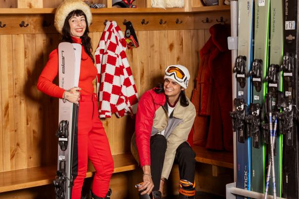 Vanessa Vanya and Alex Vigil gearing up with ski boots and skis inside the Taos Ski Valley locker room.