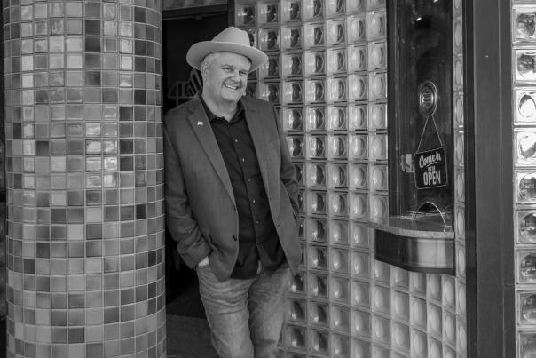 A person wearing a hat and a blazer leans casually against a tiled column outside a glass‑block‑fronted building.