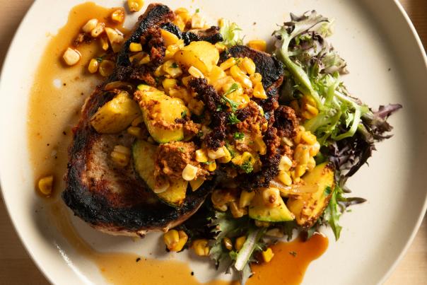 Grilled pork chop topped with roasted corn, zucchini, and crumbled sausage, accompanied by mixed greens on a beige plate.
