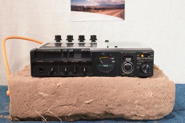 Vintage audio recorder sits on a block of adobe with a landscape photo taped to the wall behind it.