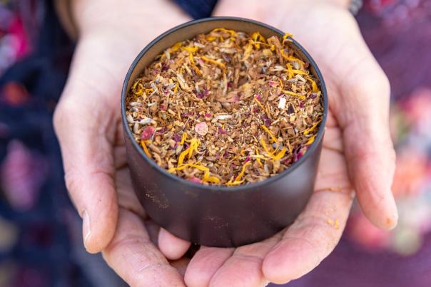 Hands gently holding a black bowl filled with an herbal blend of dried flowers and spices, including yellow petals.