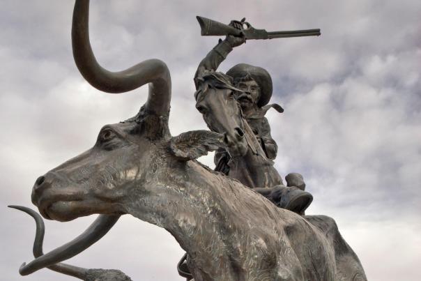 Large bronze sculpture of a rider raising a rifle while leaping forward on a charging longhorn, with additional cattle figures behind.
