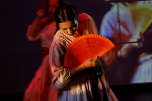 Flamenco dancers on stage holding vibrant orange fans, draped in flowing dresses, move gracefully against a dimly lit backdrop with a large projected image.