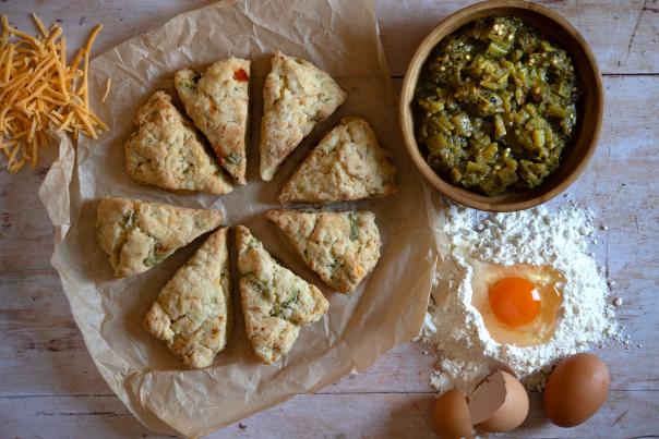A flatlay of ingredients and baked goods on a wooden surface. There is a bowl of chopped green peppers, an egg in a pile of flour, a bowl of shredded cheese, and eight baked scone triangles on parchment paper.
