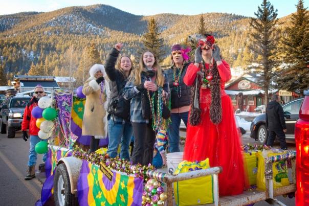 Group of people on a festive Mardi Gras float, wearing bright costumes and masks, celebrating joyfully against a snowy mountain backdrop.