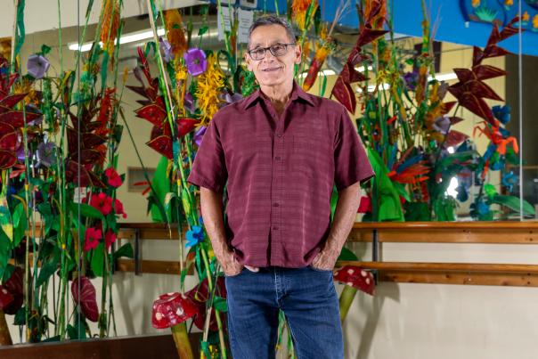 Man in a maroon shirt and jeans stands confidently in a colorful art studio with vibrant plant sculptures and large mirrors.