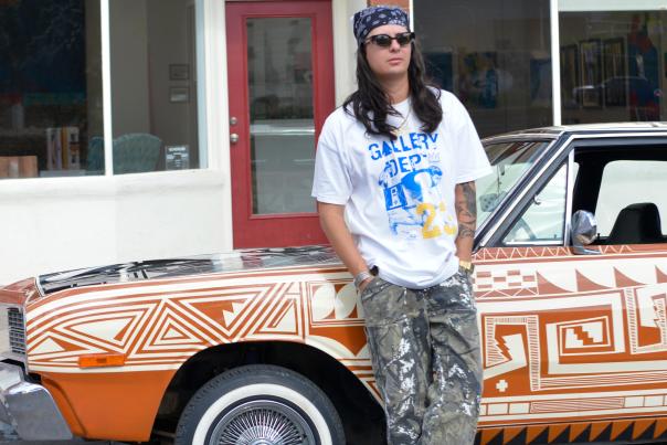 Jesse Littlebird leaning on a painted Dodge Dart outside Kukani Gallery.