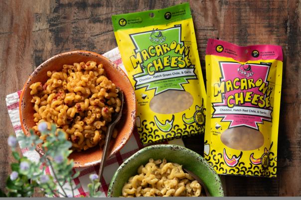 Two colorful bowls of macaroni and cheese, one orange and one green, are placed on a rustic wooden table with vibrant packaging nearby.