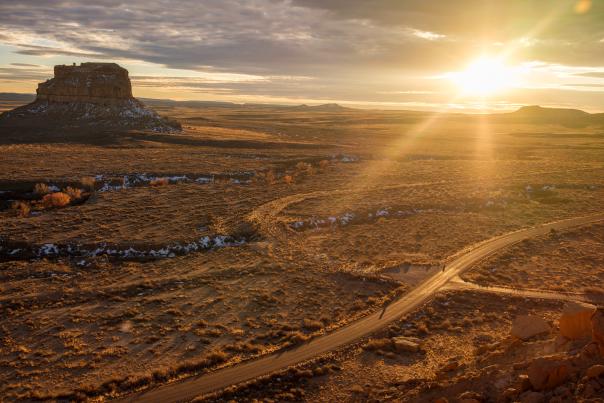 Sunset over a desert landscape with a winding dirt road, a prominent mesa, and scattered brush.