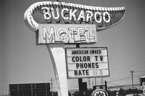 A vintage black‑and‑white roadside sign reads “Buckaroo Motel” with a silhouette of a rider atop a bucking horse above it and additional signage below advertising color TV, phones, and room rates.