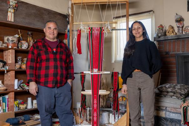 Master weaver Louie García with apprentice Allysa Concha‑Ortiz in his Albuquerque living‑room weaving studio, surrounded by yarn, tools, and an upright loom.