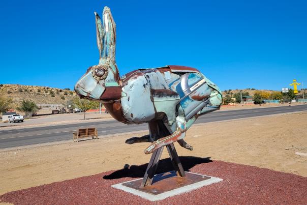 A large metal jackrabbit sculpture made from weathered, patchwork metal panels stands elevated on a wooden support beside a roadside in Gallup under a bright blue sky.