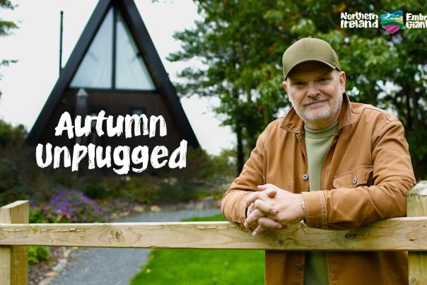 Broadcaster Ralph McLean leaning on a wooden fence in front of an A-frame cabin surrounded by trees and gardens at Ballyburren Outdoor Escapes, County Down.