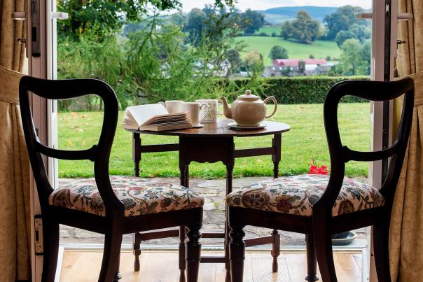 A table and chairs overlooking the scenic Clogher Valley through open French doors with a teapot, 2 cups, a milk jug and open book sitting on the table.