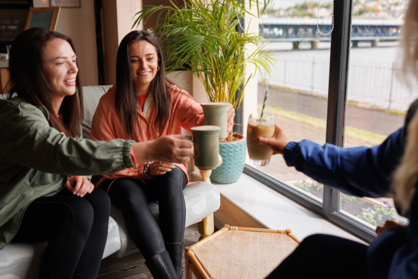 A group of women clinking their coffee mugs together with views of the River Foyle in the background.