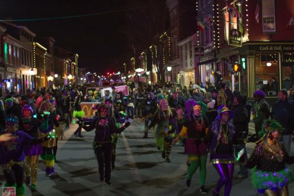 People dressed in green, purple and gold holding lights and marching in Mainstrasse Village's mardi gras parade at night.