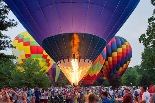 photo of large hot air balloon with fire illuminated and crowd of people and several hot air balloons in backgorund