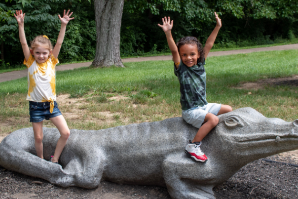 two children sitting on concrete alligator with their hands in the air, enjoying playing at Florence nature park in Florence ky