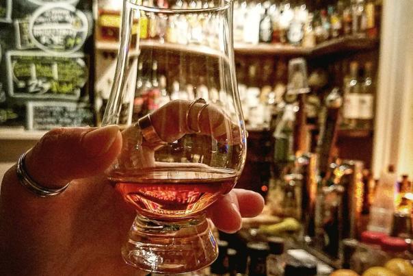 Glencairn glass of bourbon held up with Old Kentucky Bourbon Bar's huge bourbon collection behind it.