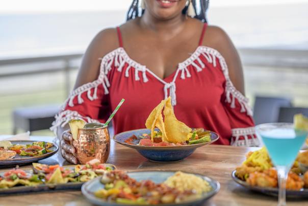 A woman in a red off-the-shoulder dress with white fringe sits at a table set with colorful dishes of seafood and cocktails. The table overlooks a coastal view, suggesting a relaxed and vibrant dining experience near the ocean.