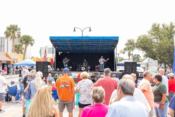 A crowd watches a live band perform on an outdoor stage at a sunny festival surrounded by palm trees and vendor tents.