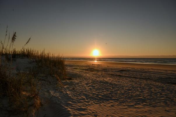 Vibrant sunrise over North Myrtle Beach, South Carolina, casting a warm, golden glow across the tranquil sandy shores. The sun's rays reflect on the gentle waves, creating a picturesque coastal scene with silhouettes of sea grass and beach dunes.