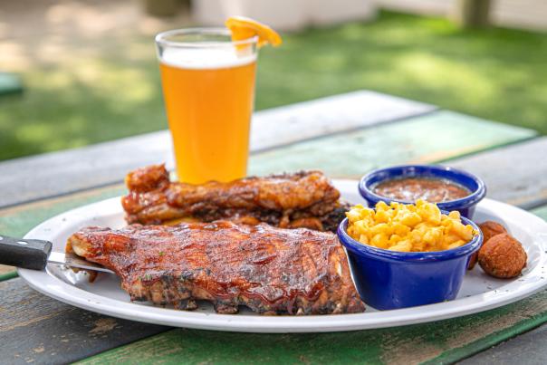 A plate of barbecue ribs served with sides of macaroni and cheese and baked beans, along with a hushpuppy. A glass of wheat beer garnished with an orange slice sits behind the plate on a weathered green picnic table in an outdoor setting, suggesting a casual and delicious Southern-style meal.