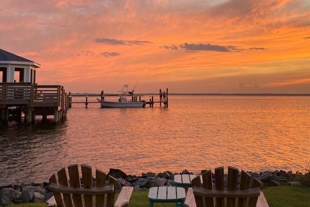 Peach and orange sunset over the sound with a gazebo over the water and addirondack chairs on the shoreline.