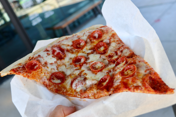 A large slice of pepperoni pizza with melted cheese is held on a sheet of white parchment paper outside Atomic Cowboy in Downtown Overland Park, with outdoor seating and a glass door visible in the background.