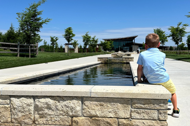 A young boy sits on the edge of the fountains in front of Longhouse Visitor's Center at the OP Arboretum on a sunny day. The path is lined with green grass, trees, and a wooden fence under a clear blue sky.