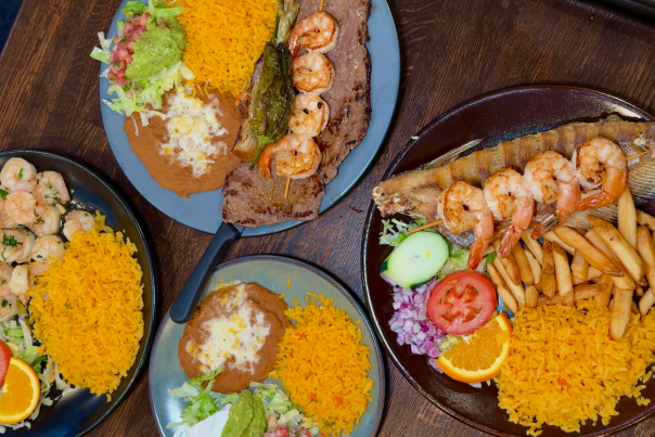 Overhead view of four plates featuring Mexican seafood dishes, including grilled shrimp served over grilled steak and whole fried fish, accompanied by orange rice, refried beans with cheese, lettuce, pico de gallo, and garnishes like orange slices, tomato, and cucumber. One plate features shrimp sautéed with herbs, served with rice and salad.