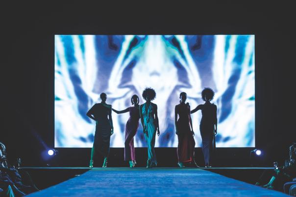 The silhouette of five women standing on the runway in front of a large screen.