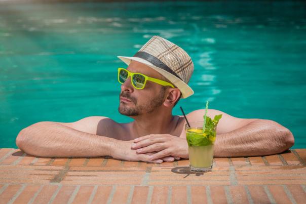 Portrait of a handsome man resting at the edge of a swimming pool. Model is wearing hat and sunglasses. mojito cocktail next to him. Summer vacation concept.