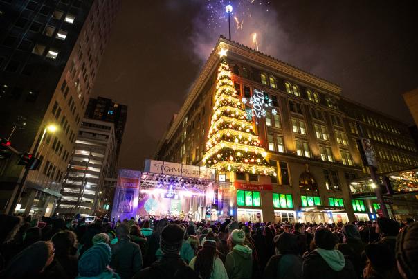 A lit Christmas tree display on the corner of a building and a stage below it with a crowd of people watching.