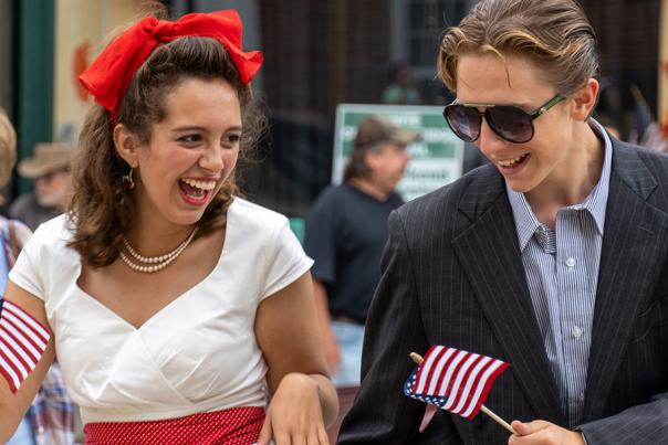 Girl with a bright red bow and pearls dancing with a man in a suit and sunglasses on a street with other people.