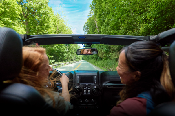 Two women in a corvette driving down an open road with green trees framing the road