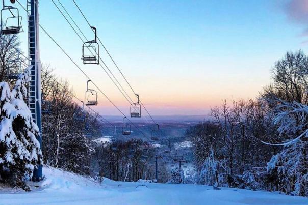 Sunset view from a ski mountain with trees along either side of the ski slope and a ski lift in frame.