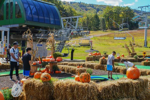 A man playing mini golf with three children. The mini golf course is decorated with pumpkins and hay bales.