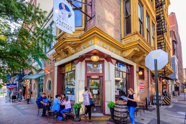 The exterior of a storefront on the corner of a street intersection with people sitting outside and walking in the store