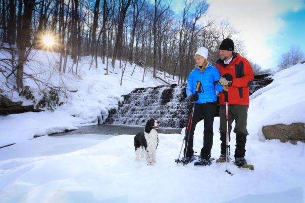 A man and a woman in winter attire snowshoeing with a dog accompanying them