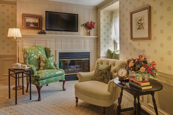 Two sitting chairs with side tables in front of a a fireplace with a television hanging overhead.