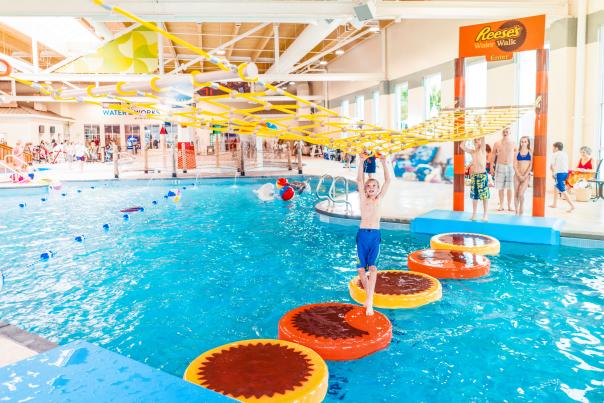 Young boy in a swim suit crossing floating pads using a net that hangs above him at Hershey Lodge's indoor waterpark.