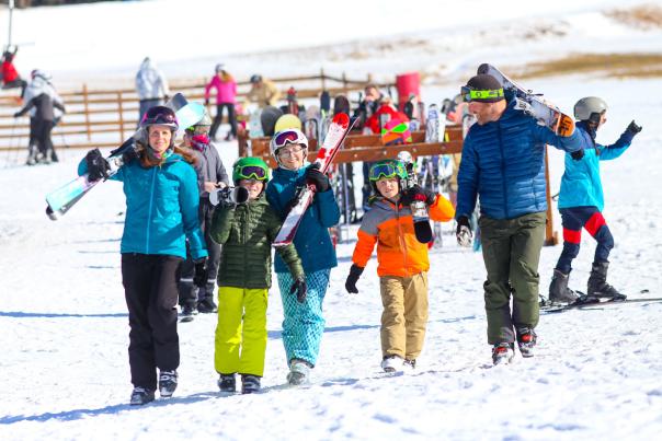 Two adults and three children in winter gear and carrying skis in their arms walking at a ski resort.