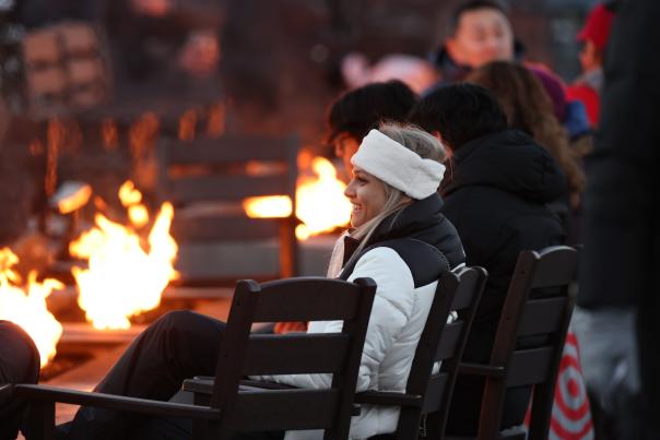 Woman in snow gear sitting around an outdoor firepit with other people.